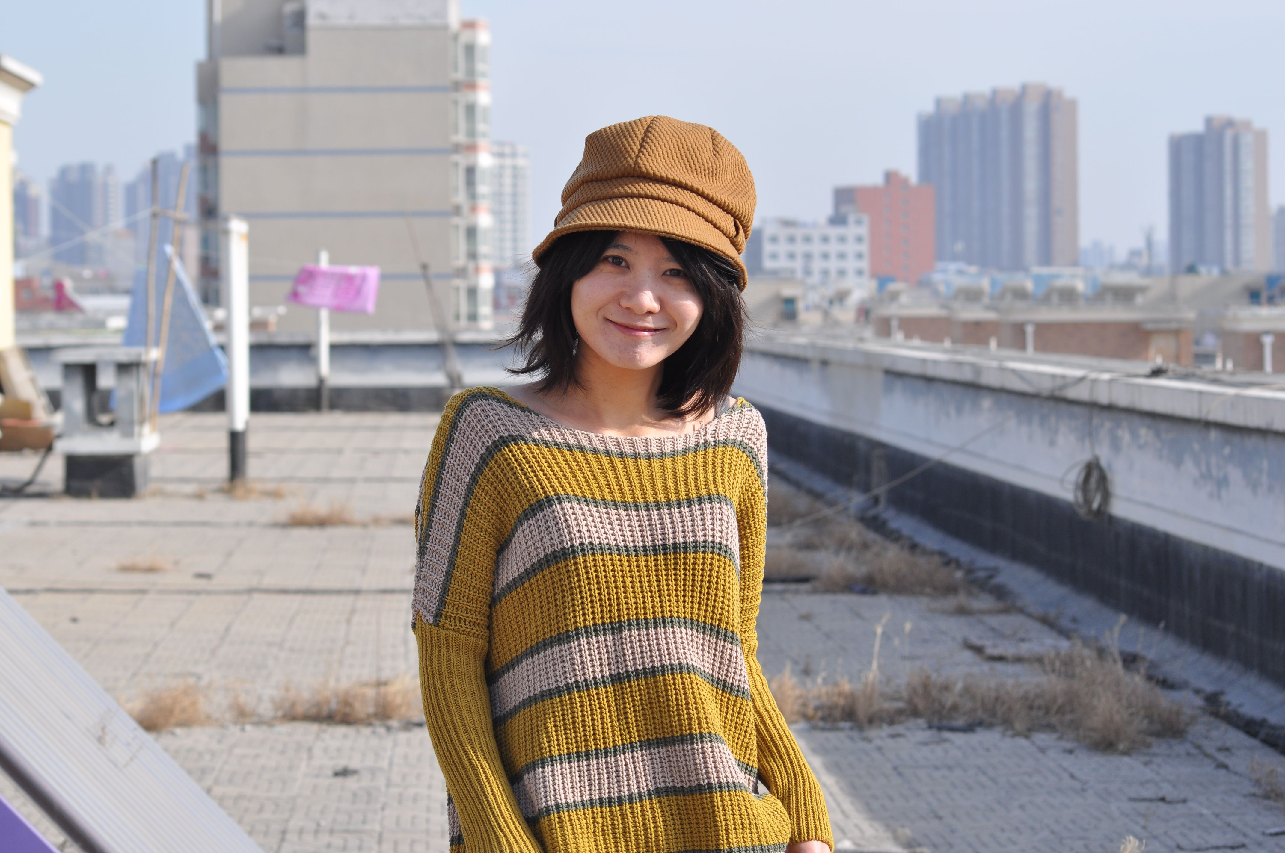 Woman in striped sweater and hat smiles on an urban rooftop with cityscape background.