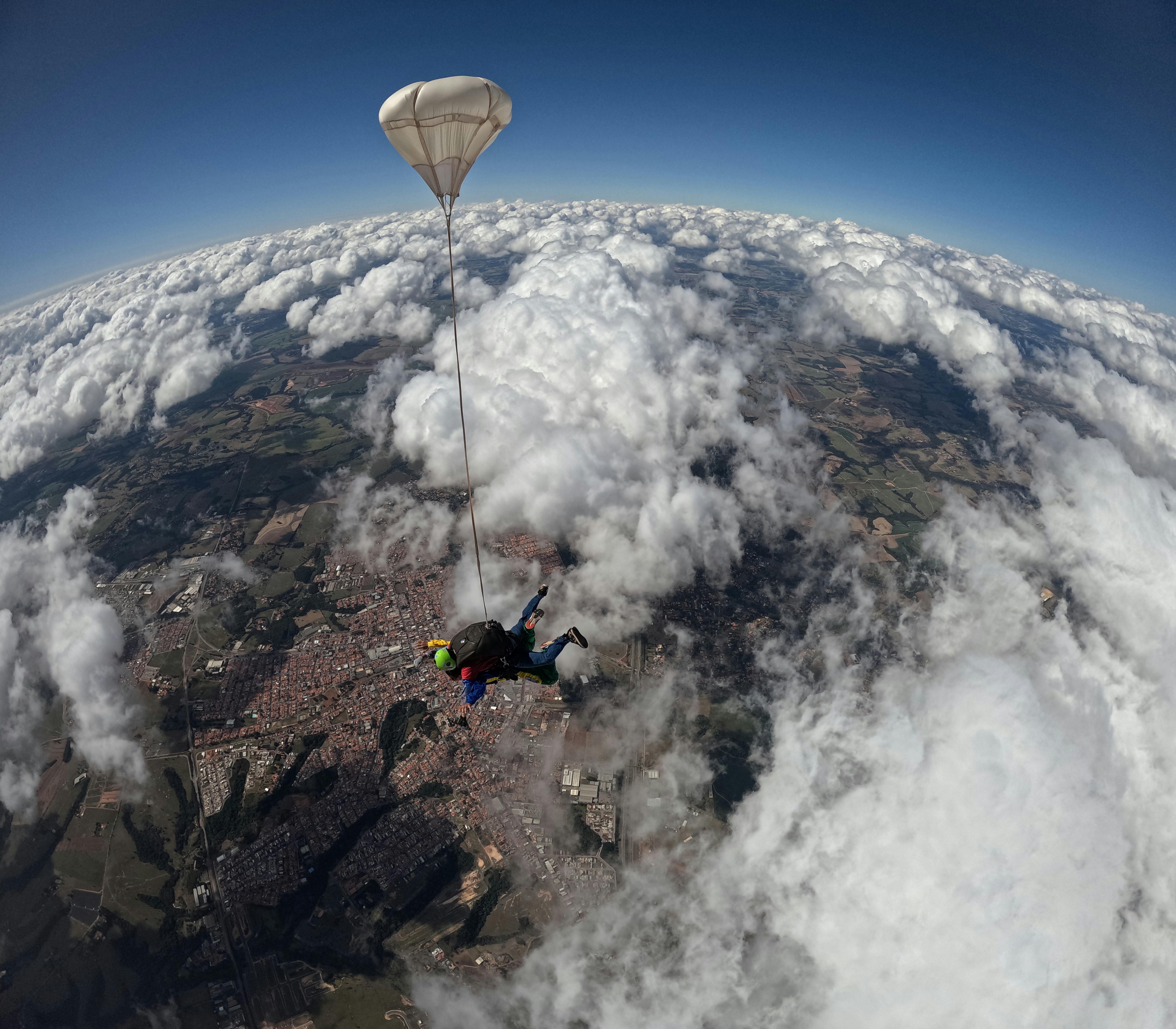 A thrilling aerial shot of a skydiver under parachute above a city and clouds.