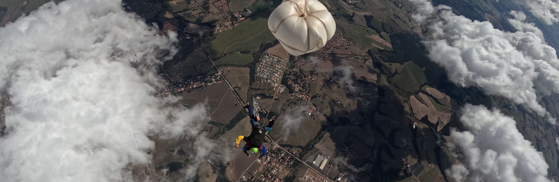 Skydiver descending with parachute over a vast landscape, surrounded by clouds.