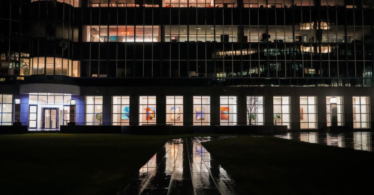 Illuminated office building exterior with reflections on wet pavement at night.
