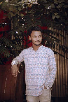 Young man in traditional attire standing outdoors, surrounded by lush green foliage.