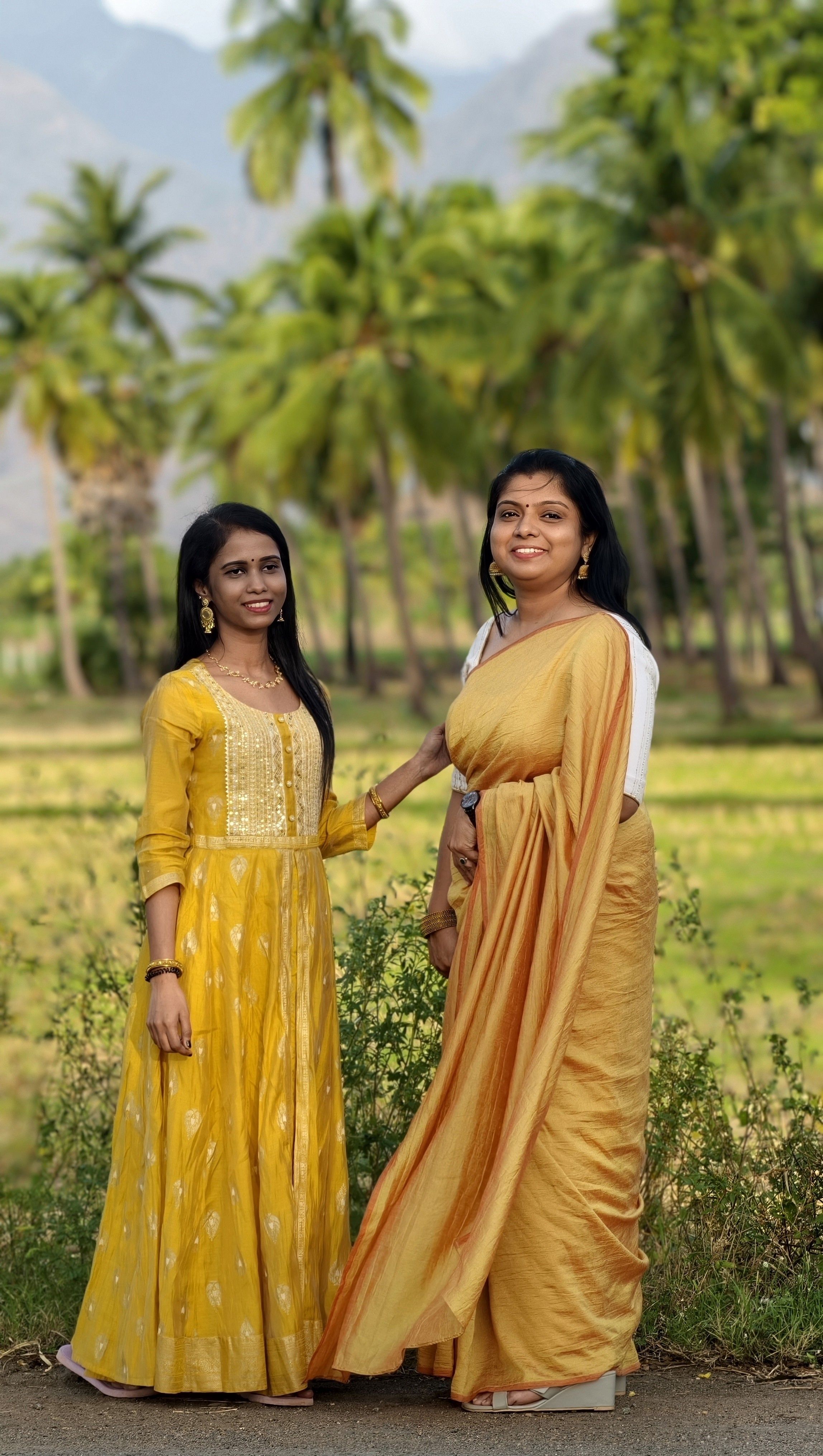Mother and daughter pose in vibrant traditional dresses amidst lush palm trees in rural Tamil Nadu, India.