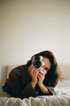 A woman lay on bed capturing a selfie with a classic film camera.