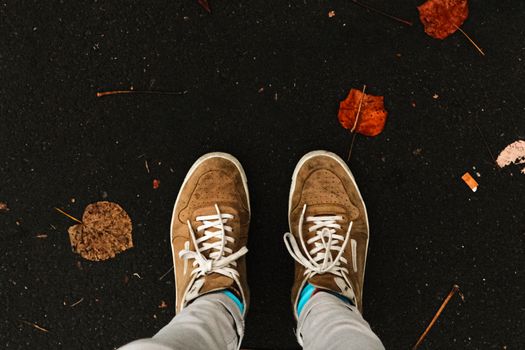 Brown sneakers on a dark pavement with scattered autumn leaves, embodying a fall vibe.