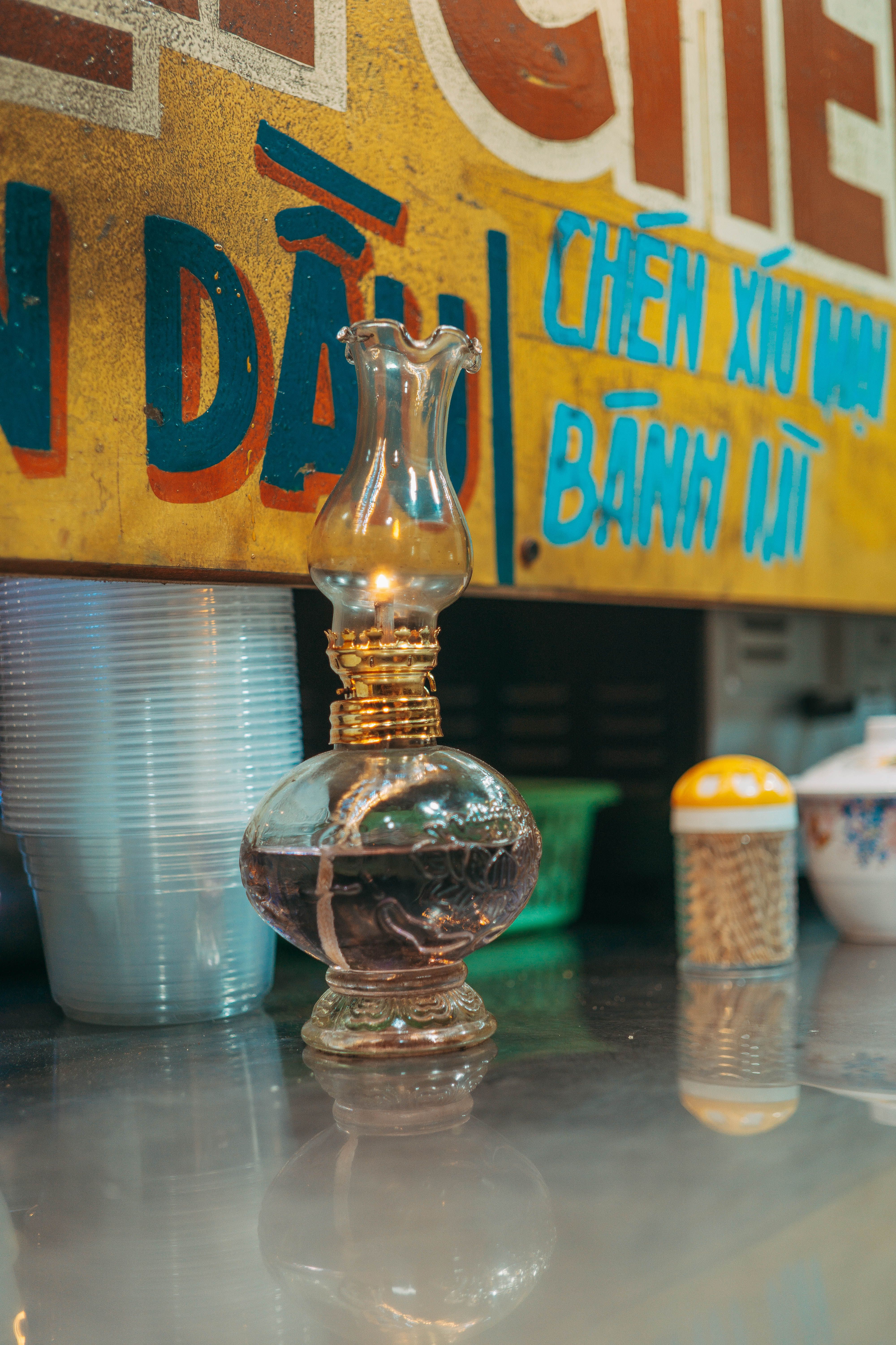 A vintage oil lamp on a reflective surface with colorful Vietnamese signage in the background.