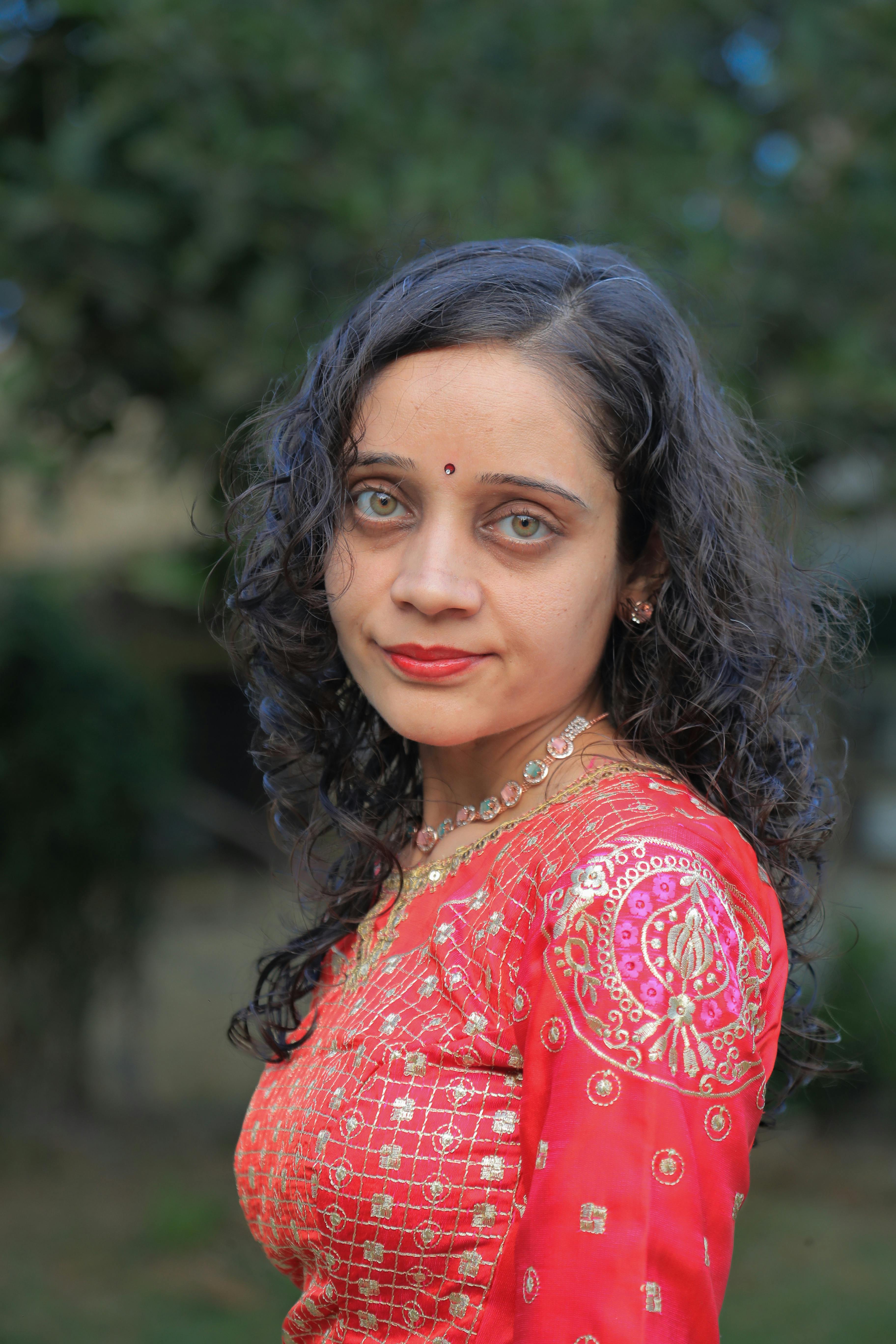 Portrait of a South Asian woman in traditional red attire, standing outdoors with a nature backdrop.