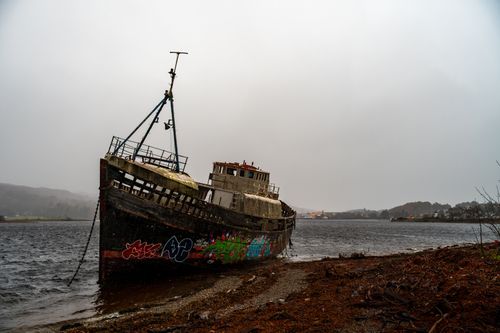 Free A derelict ship adorned with graffiti rests beside a tranquil lake under cloudy skies. Stock Photo