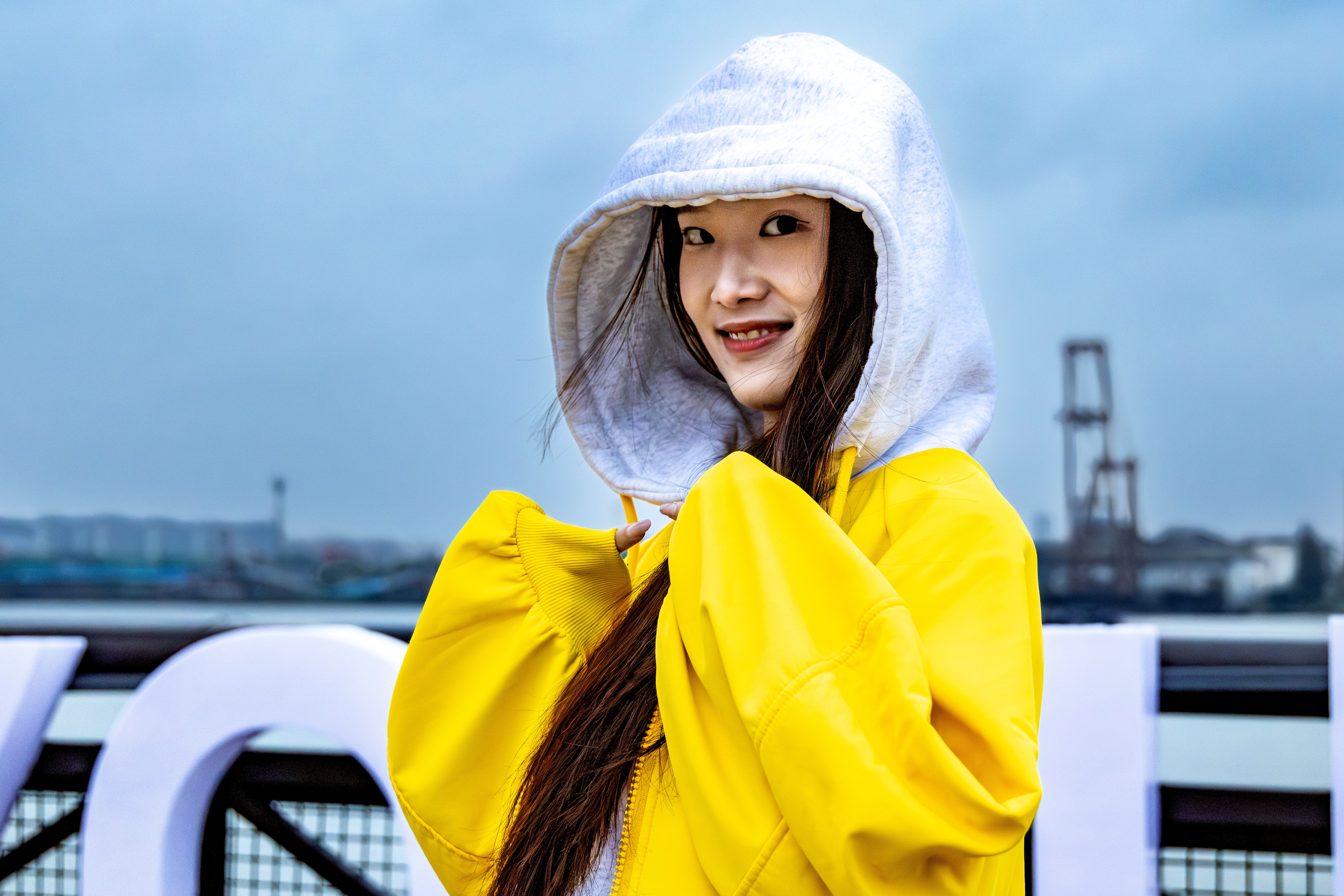 Smiling woman in yellow jacket with hood up, standing by a harbor on a cloudy day.