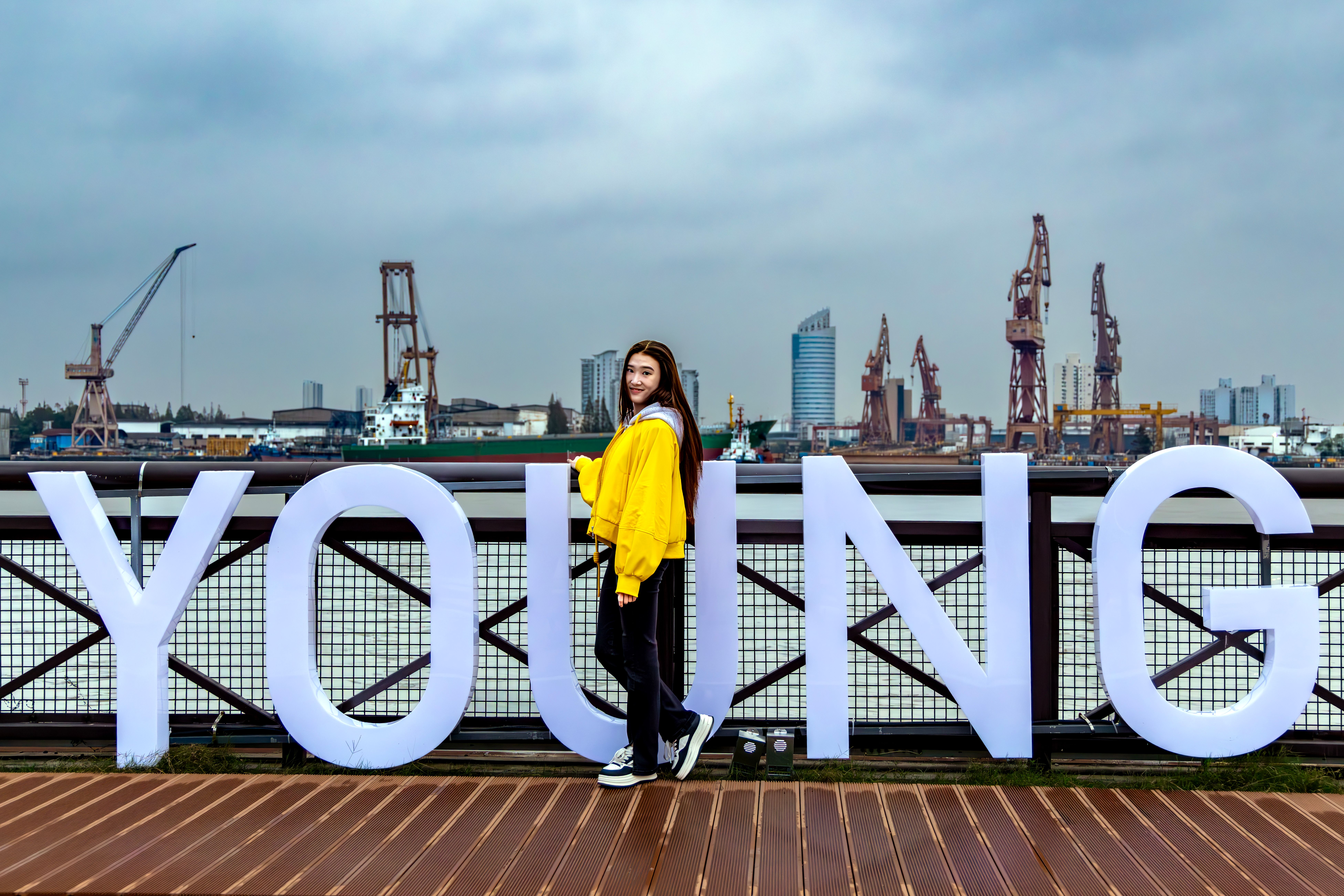 A young woman poses next to large letters spelling 'YOUNG' with an industrial waterfront backdrop.