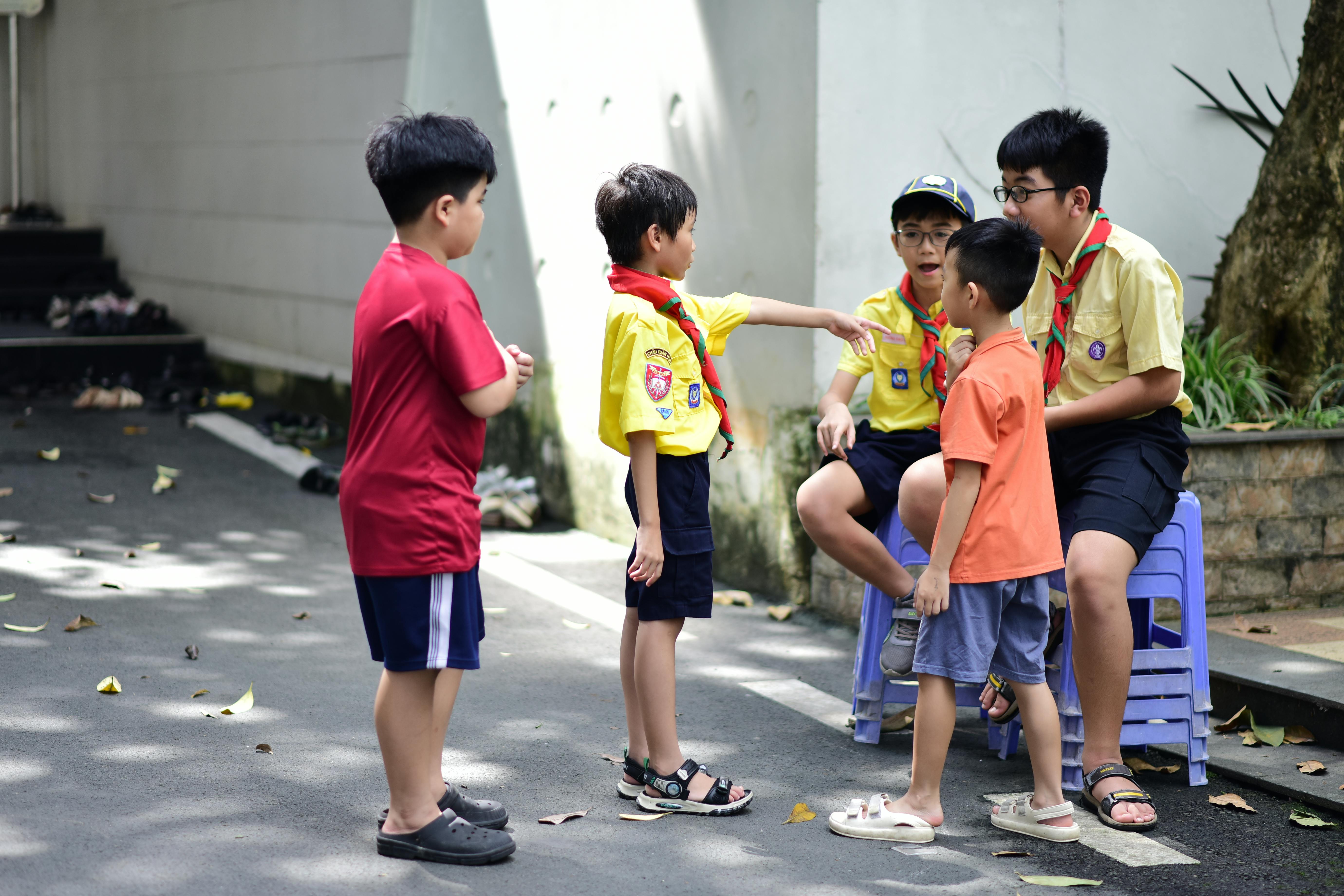 Five children in scout uniforms interacting outdoors on a sunny day.