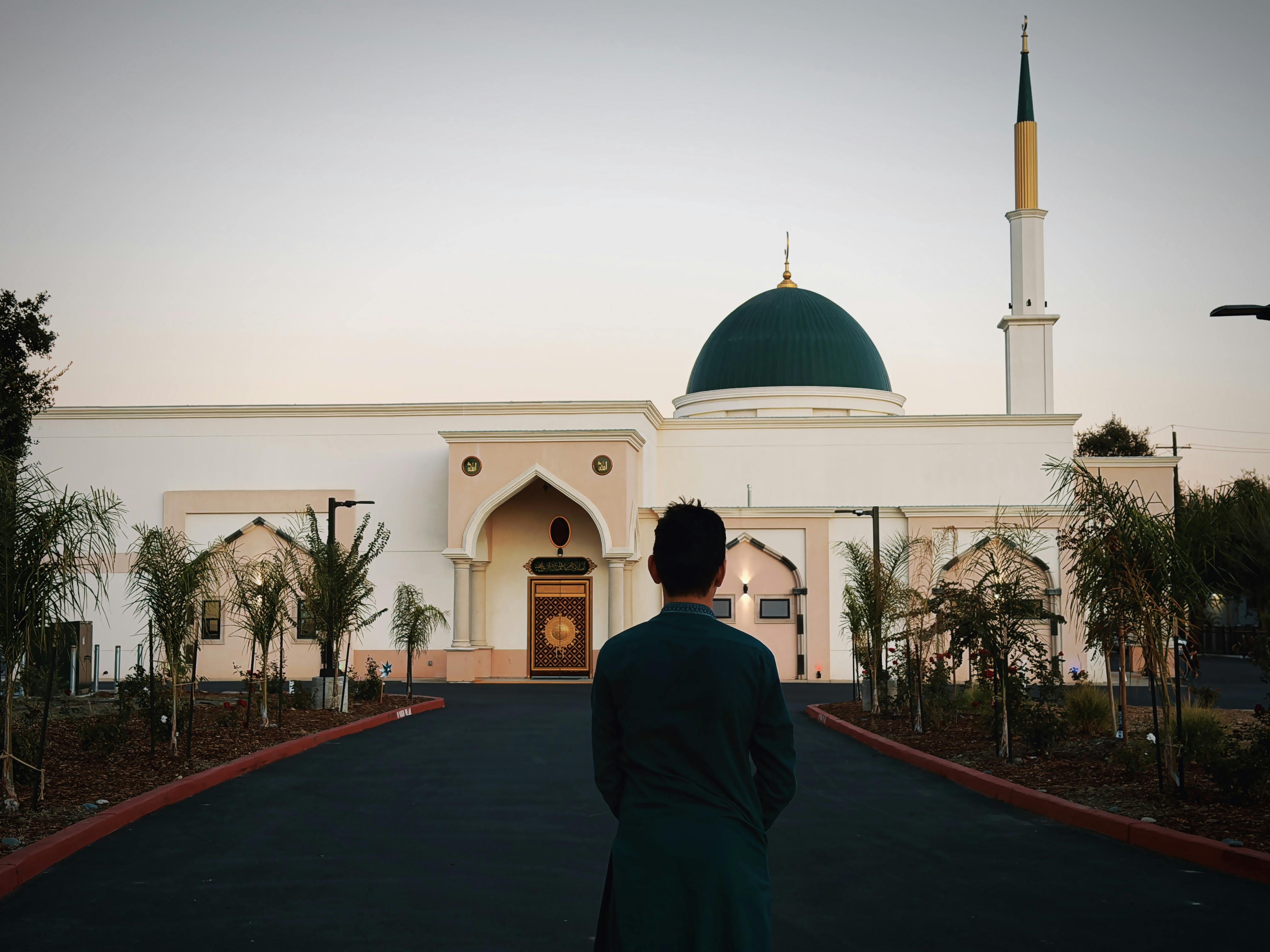 A man stands outside a beautifully designed mosque with a green dome at sunset.