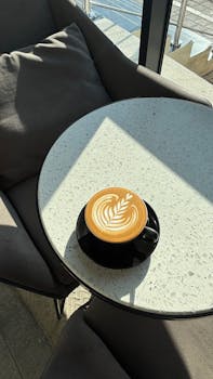 Latte art in a black cup on a sunlit table by a window in a modern cafe.