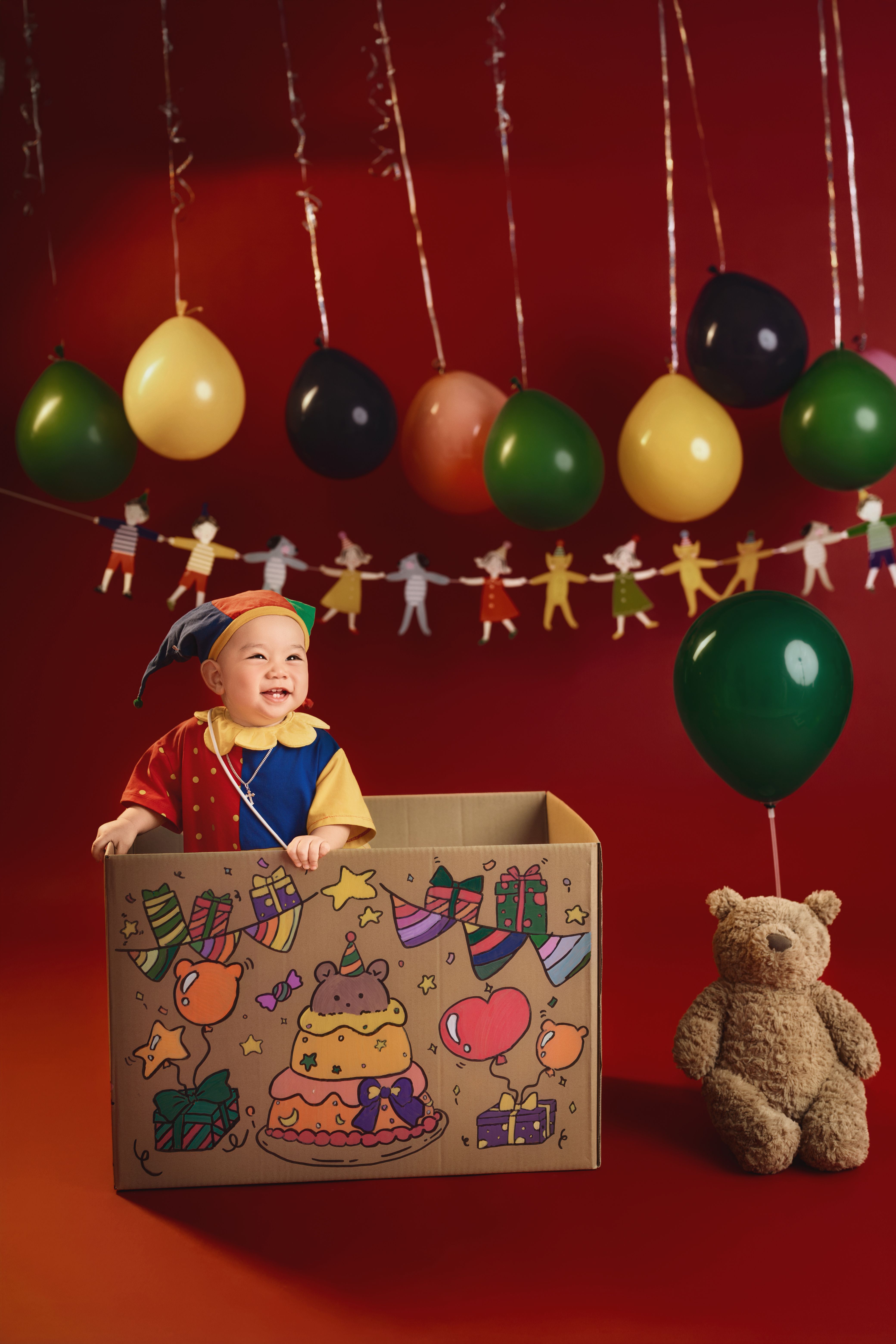 Joyful baby in a colorful cardboard box with balloons and teddy bear in celebration decor.