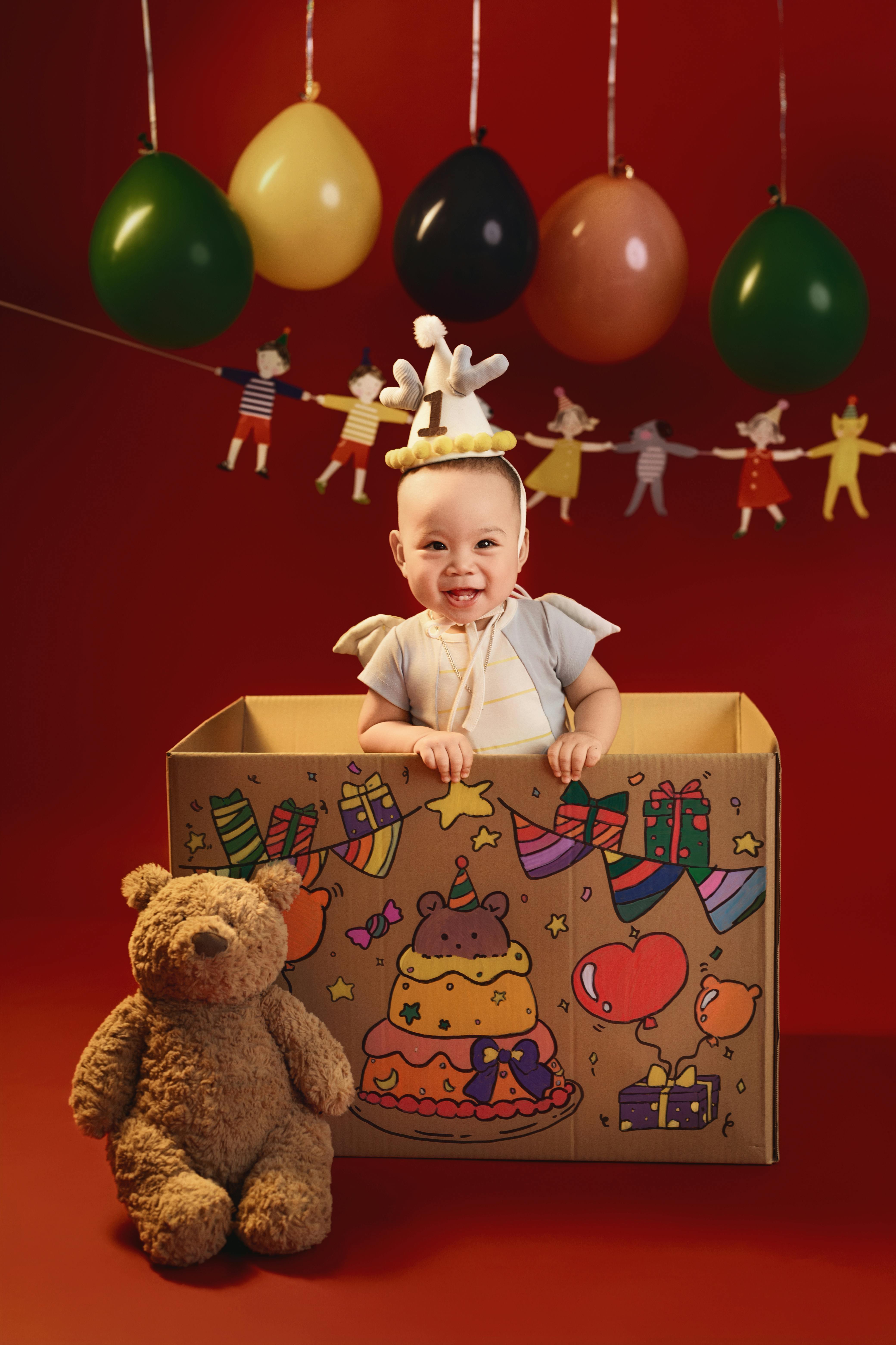Happy baby in a decorated box with teddy bear and balloons, perfect for birthday themes.