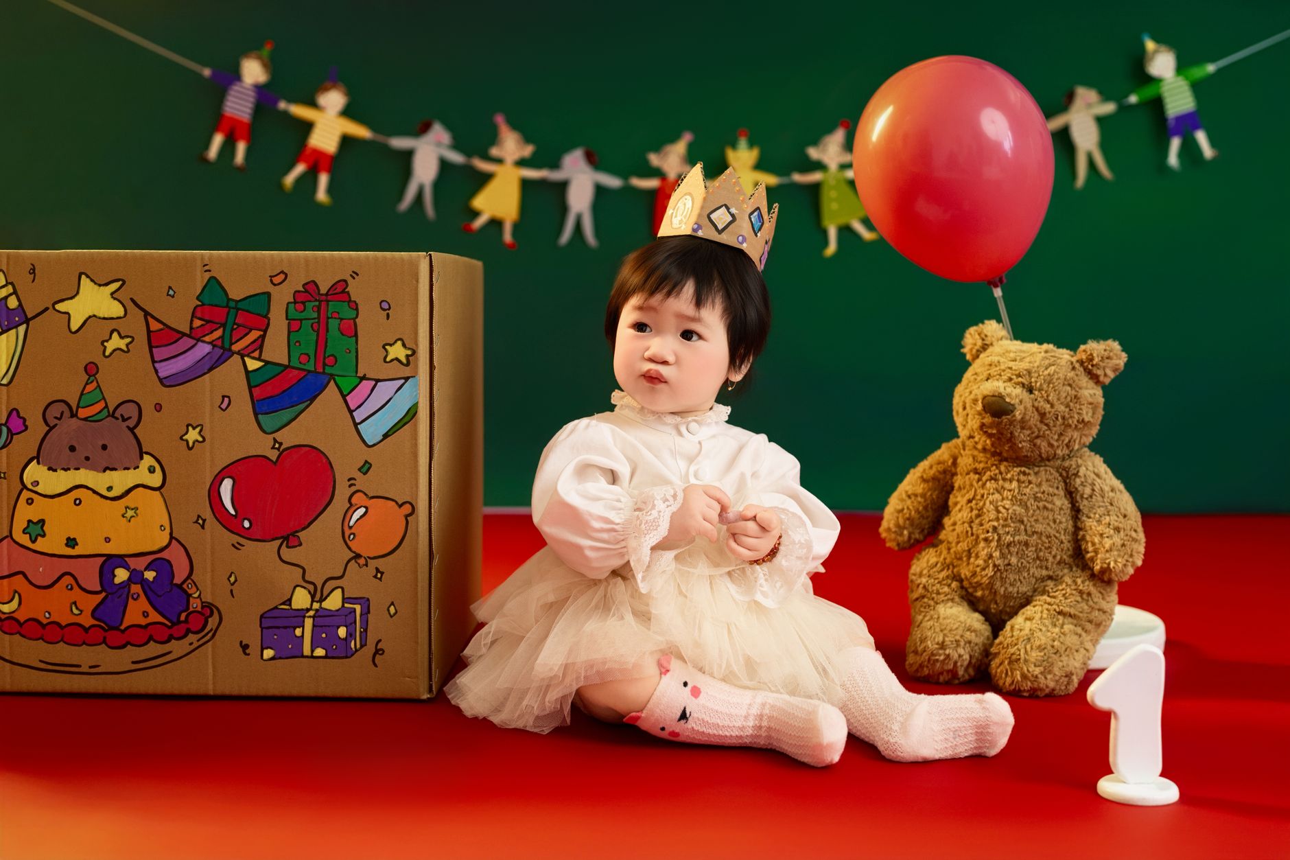Cute baby in white dress and crown celebrating first birthday with teddy and balloons in a festive setup.