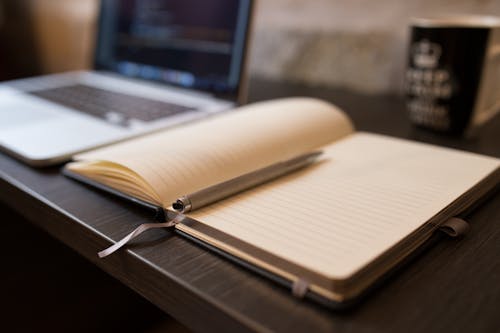 Grey Click Pen on Opened White Lined Book Beside Opened Macbook Pro on Table