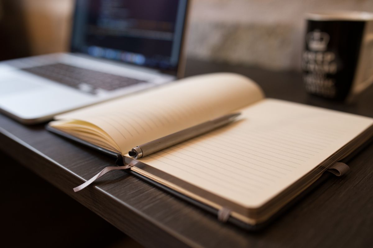 Open notebook with pen beside a laptop on a wooden desk, ideal for office or study themes.