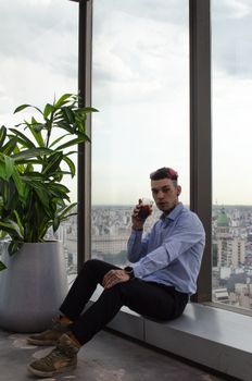 Man in blue shirt sits by a tall window with city skyline view, holding a drink.