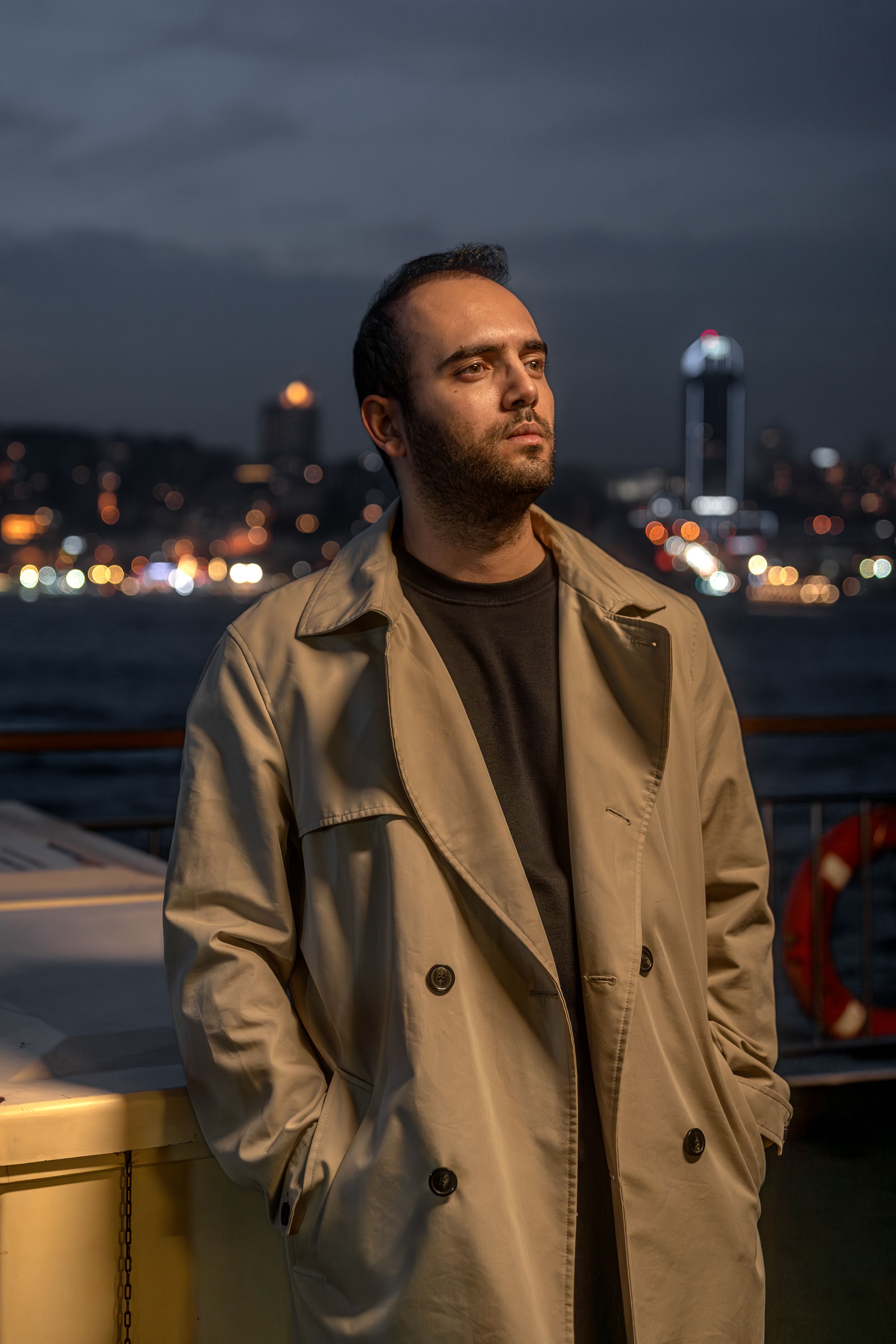 Man in trench coat stands on a ferry at night with city lights in the background.