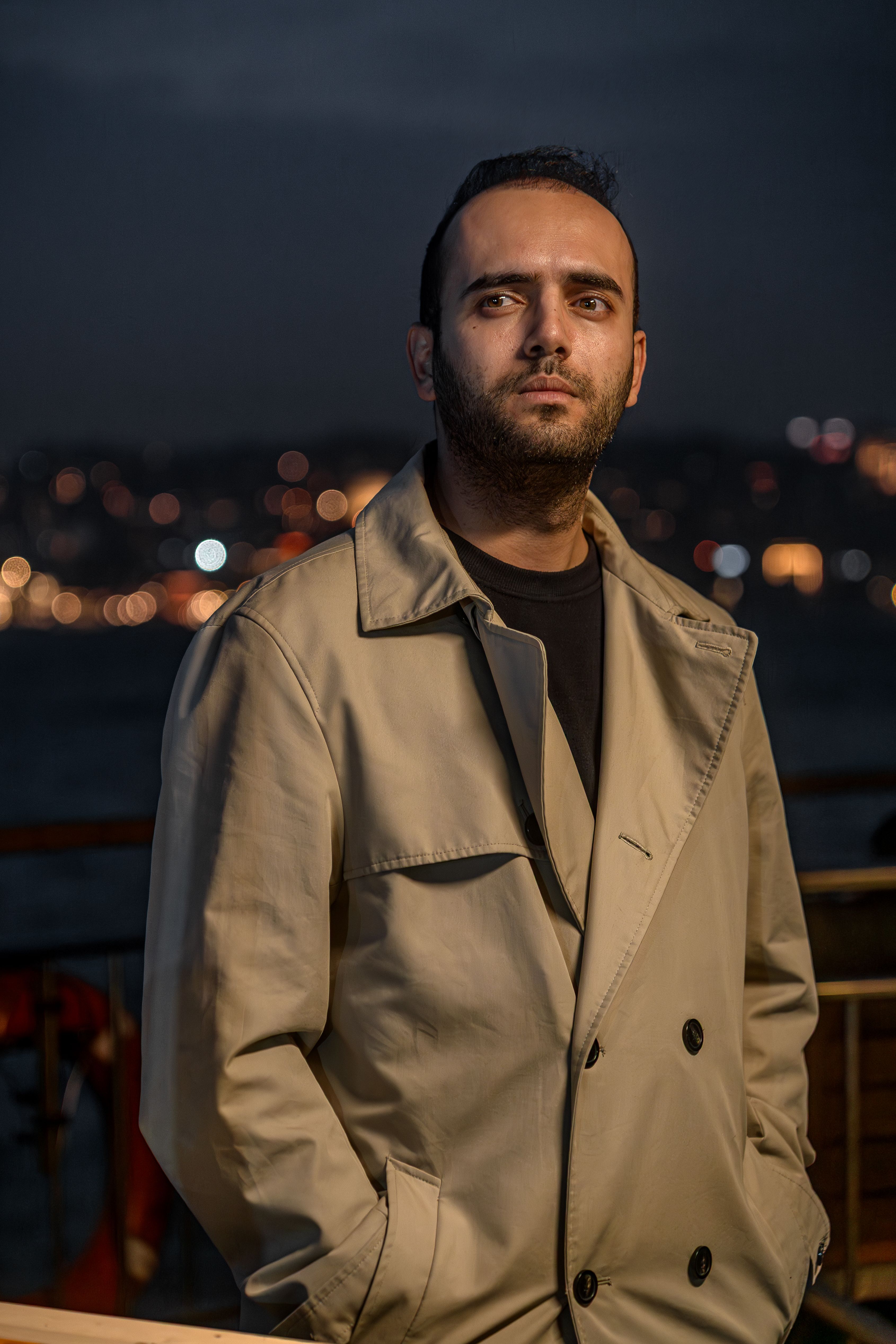 Portrait of a man in a beige trench coat with city lights in the background at night.