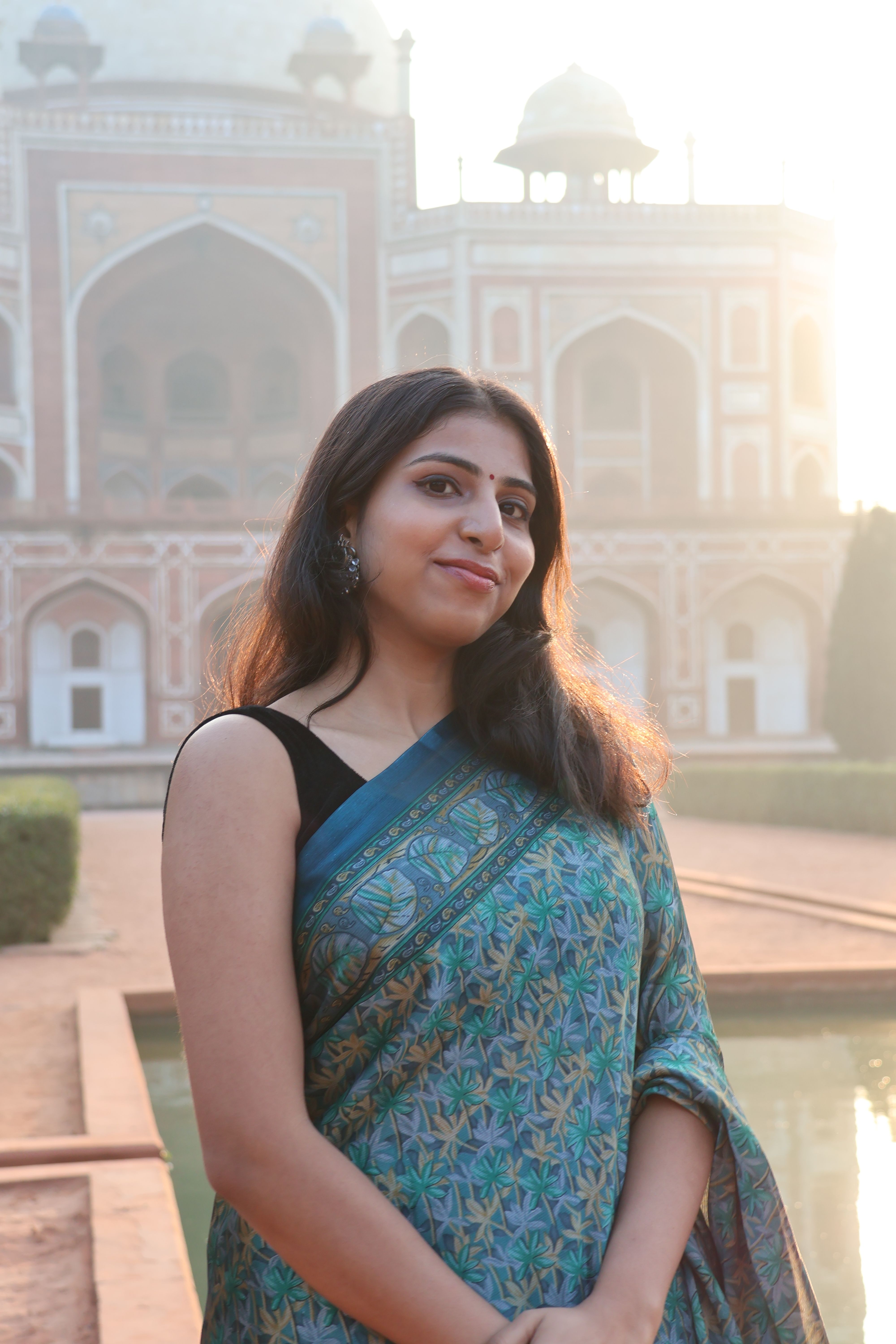 A woman in a blue saree poses in front of Humayun's Tomb in Delhi, bathed in warm sunlight.