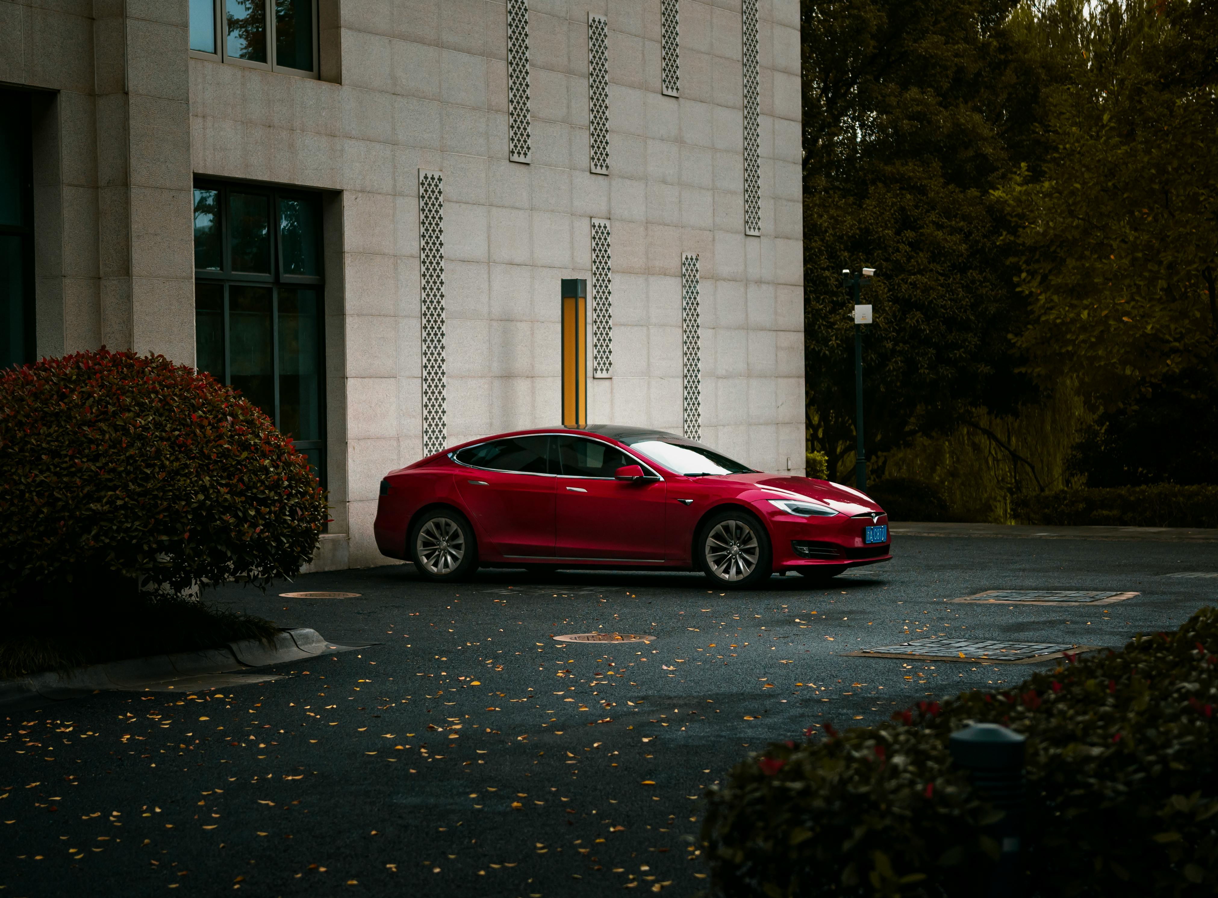A stylish red electric car parked beside a modern building with landscaping and evening lighting.
