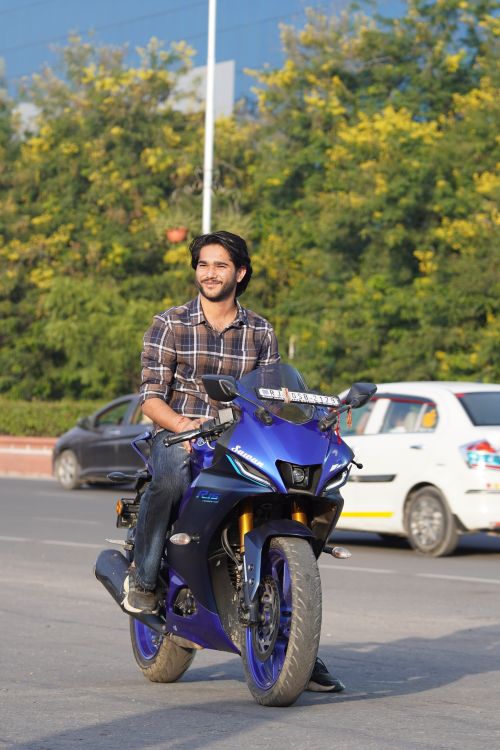 Free A young man rides a blue motorcycle on a sunny day with cars and greenery in the background. Stock Photo