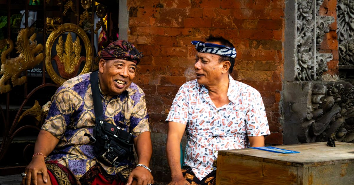 Two Indonesian men smiling and laughing while sitting outdoors in traditional clothing.