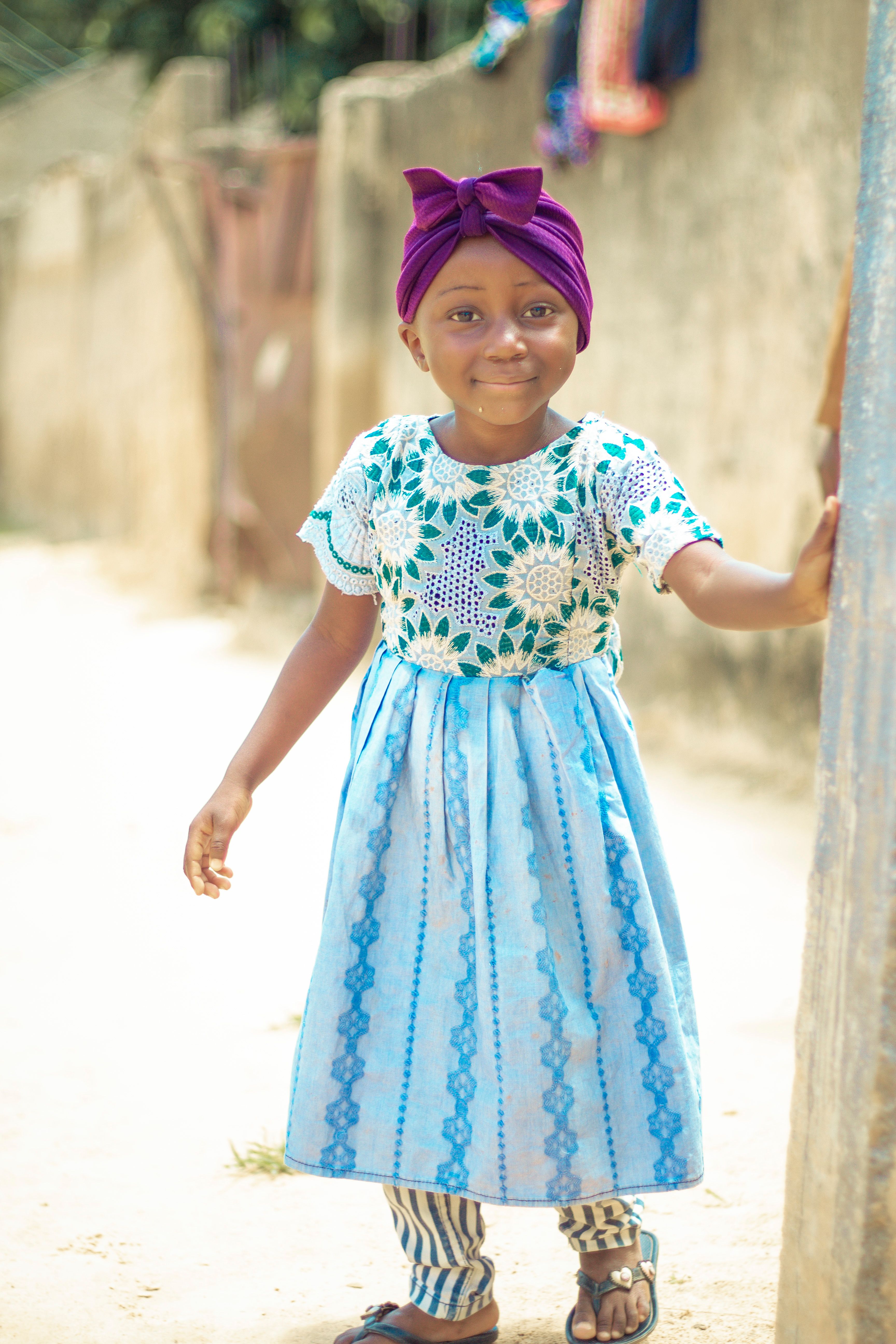 A young girl wearing a blue dress and headband smiles outdoors in Yobe, Nigeria.