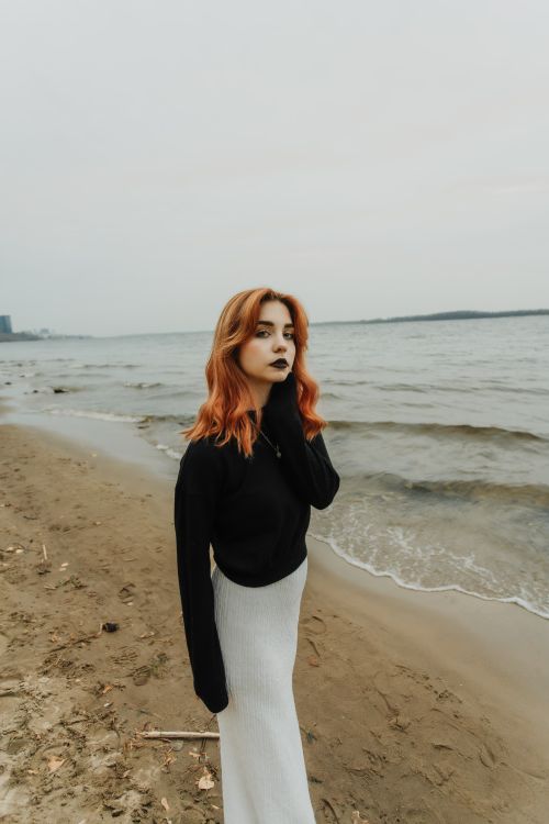 Free Woman with red hair on a sandy beach wearing a black sweater and white skirt facing the camera. Stock Photo
