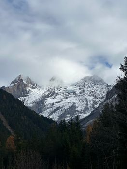 Snow-capped peaks of a mountain range under a cloudy sky, with lush forest at the base.