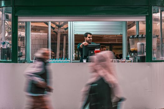 Street vendor serving drinks interacts with passing pedestrians in an urban setting.
