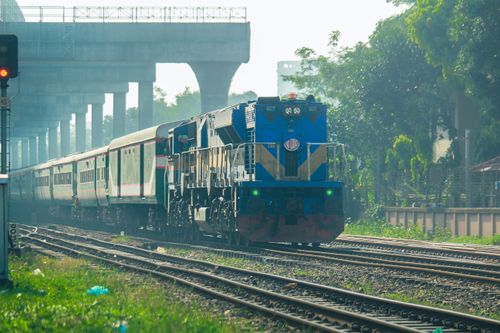 Free A blue train travels through a lush urban area under an overpass on a sunny day. Stock Photo