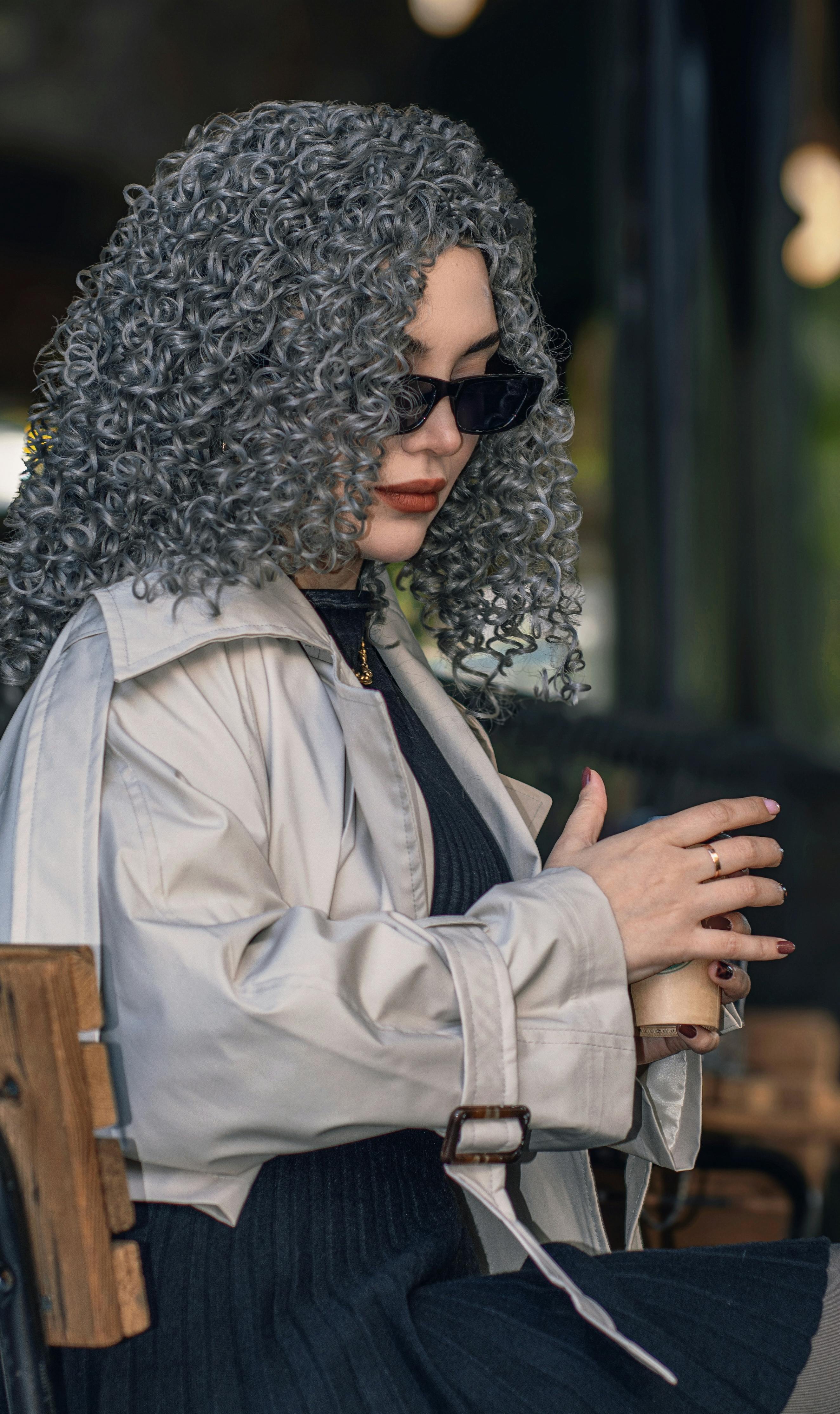 Gratis Donna elegante con capelli grigi e ricci e occhiali da sole che si gode un caffè all'aperto. Abbigliamento autunnale alla moda. Foto a disposizione