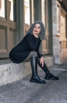 Fashionable woman with curly gray hair seated outdoors in city.