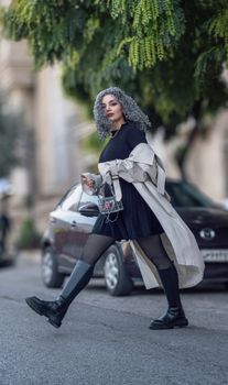 Fashionable woman in urban setting with gray hair, wearing a trench coat and boots, walking confidently.