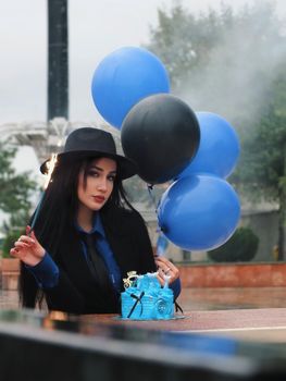 Elegant woman holds blue and black balloons, celebrating with a cake outdoors.