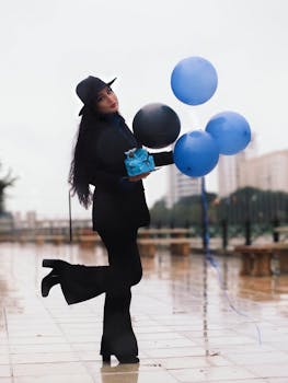 Fashionable woman in black holding blue balloons and gift on a wet urban street.