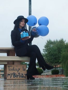 Woman in black suit and hat celebrates birthday outdoors with blue balloons and a cake.