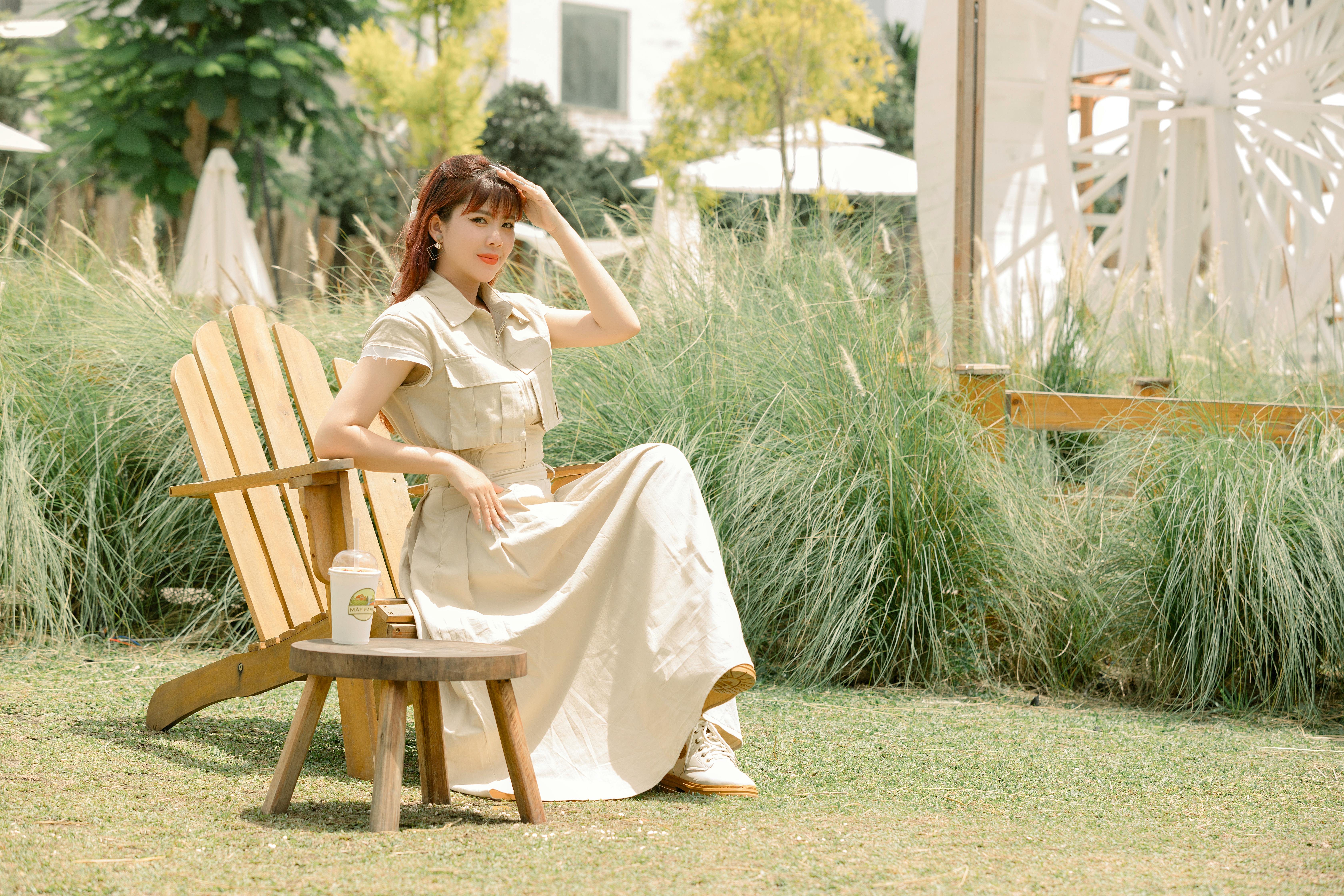 A woman in a light dress sits on a wooden chair in a sunny garden setting.