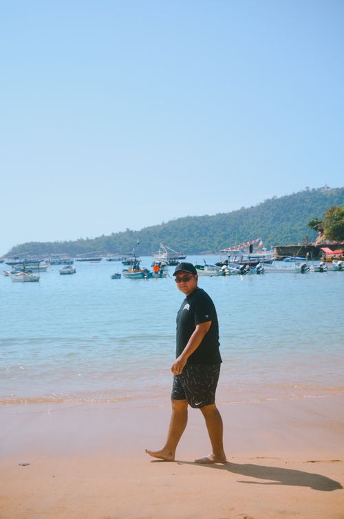 Free A man strolls along a sunny beach with boats anchored offshore under a clear blue sky. Stock Photo