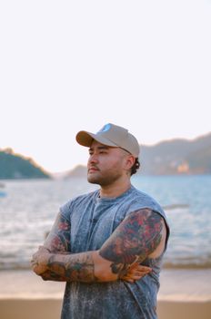 A tattooed man in a cap standing confidently by the sea with mountains in the background.