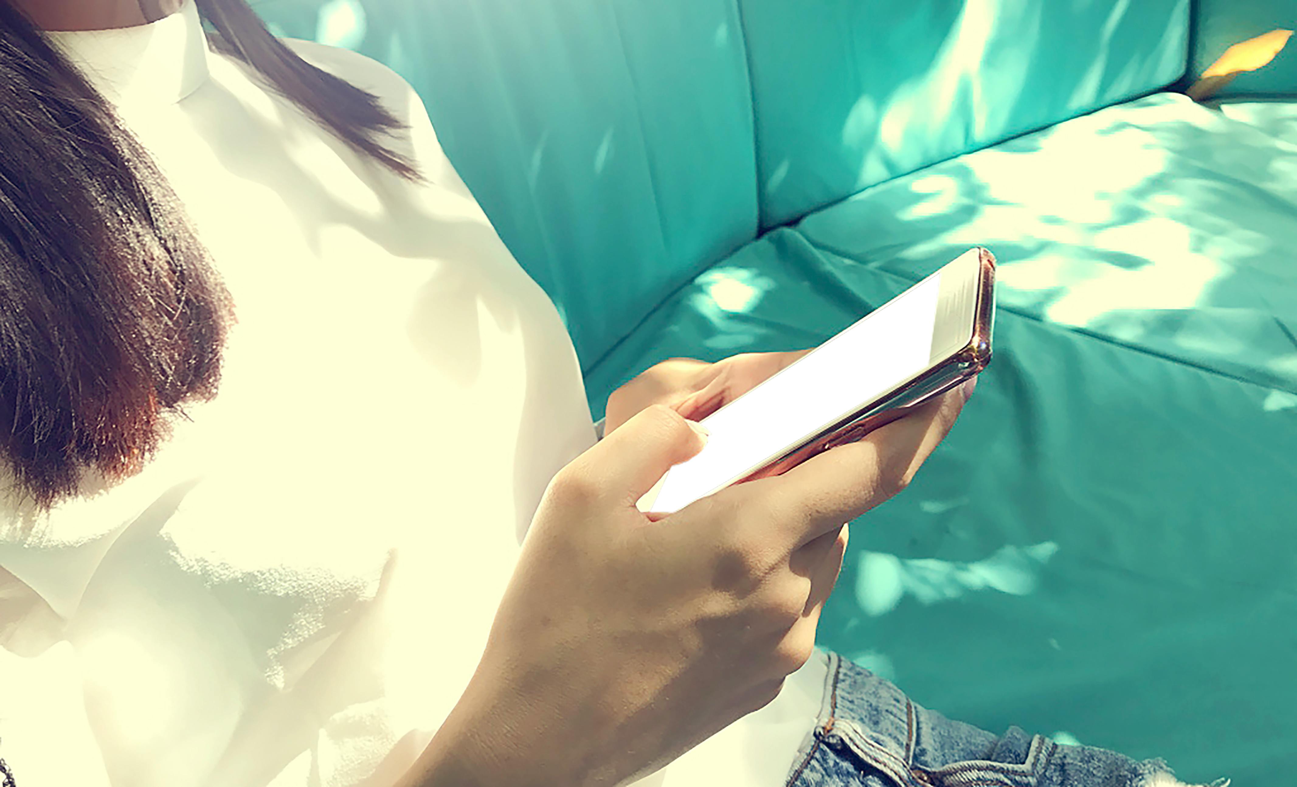 一位穿着休闲装的女人坐在阳光斑驳的沙发上用着智能手机