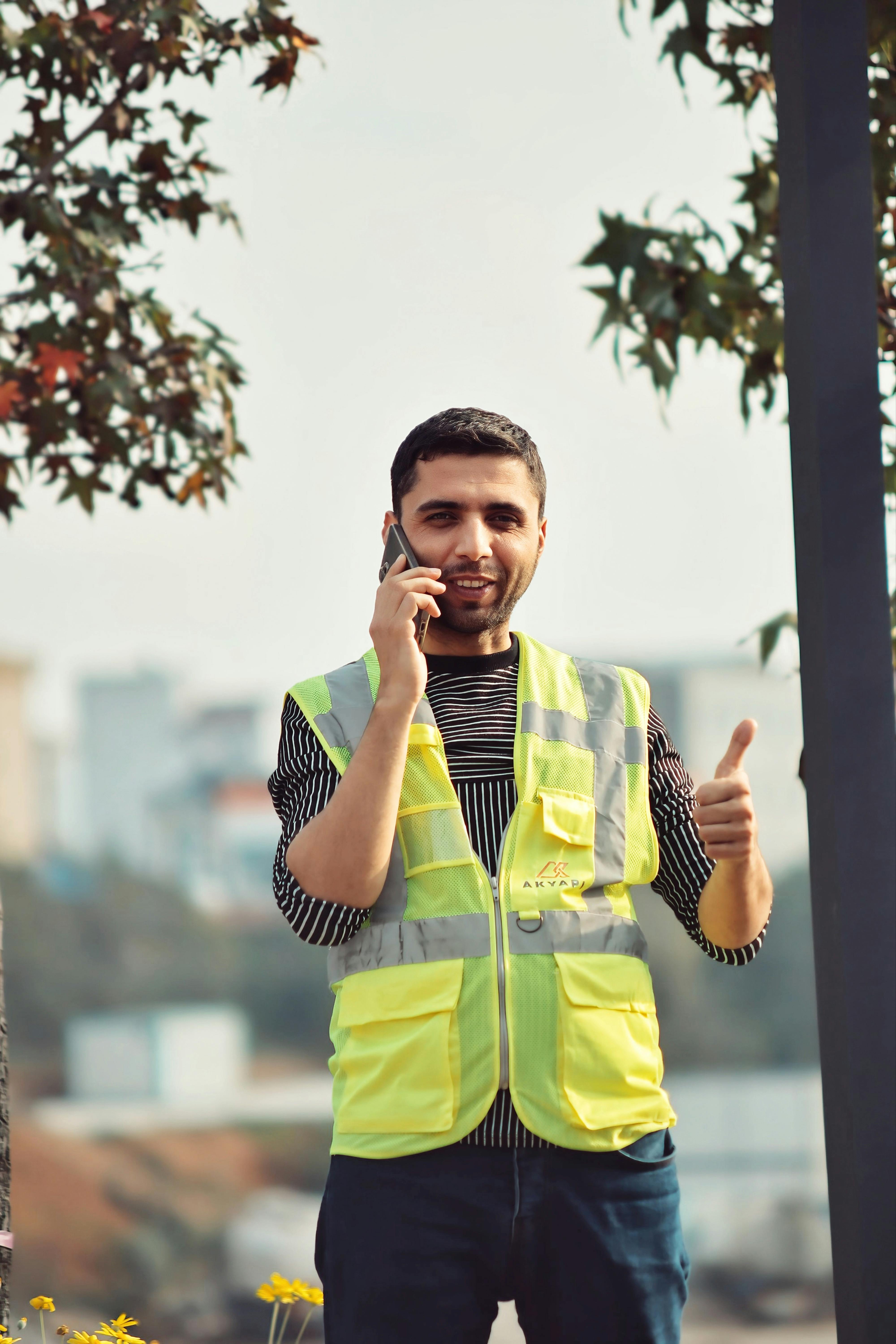 Young construction worker in a reflective vest talks on the phone and gives a thumbs up outdoors.