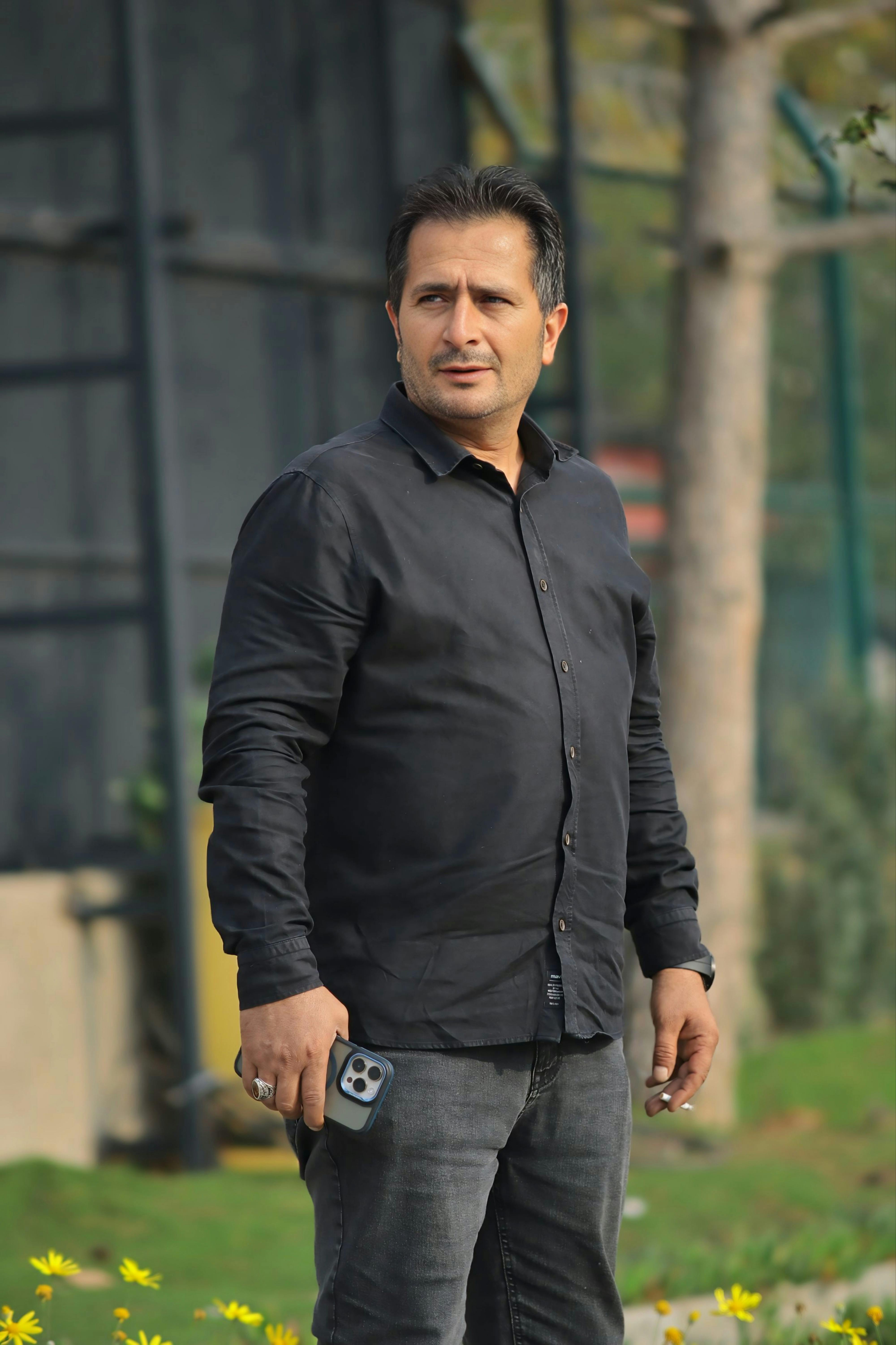 Man in a black shirt holding a smartphone outdoors, looking away, natural setting.