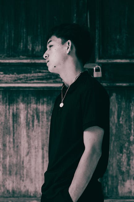 Side profile of a young man standing against a textured wall, wearing a necklace.