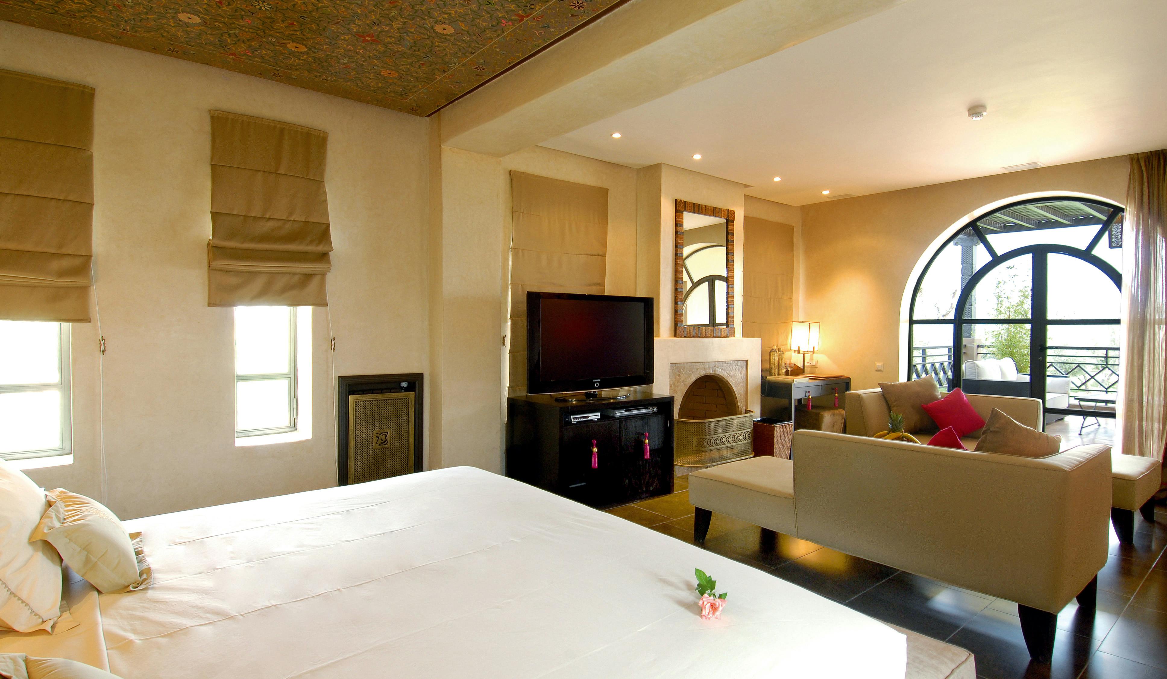 Free Elegant modern bedroom suite with cozy seating, large windows, TV, and fireplace, exuding warmth and luxury. Stock Photo