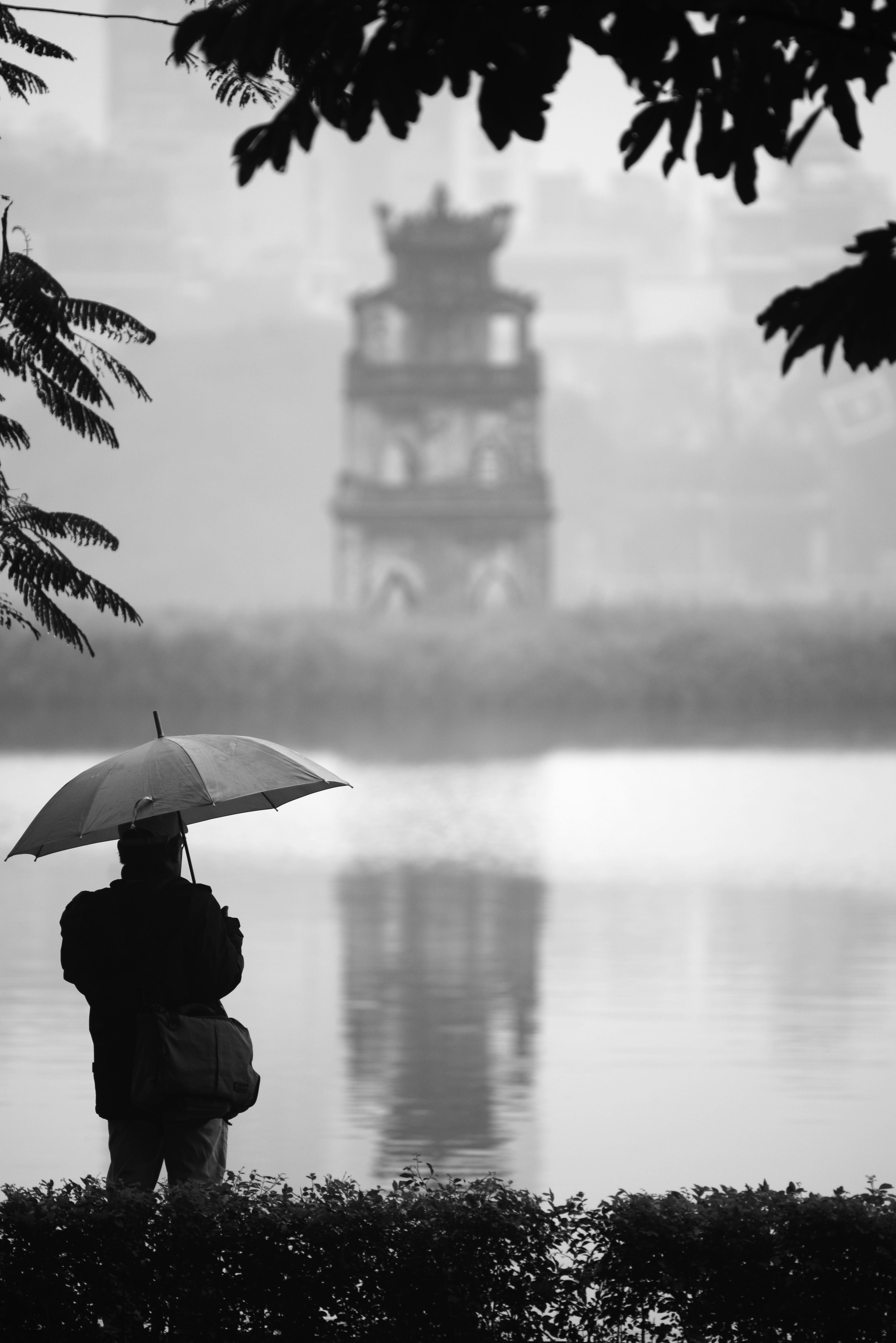A contemplative silhouette with umbrella at Hoan Kiem Lake in a rainy, misty setting.