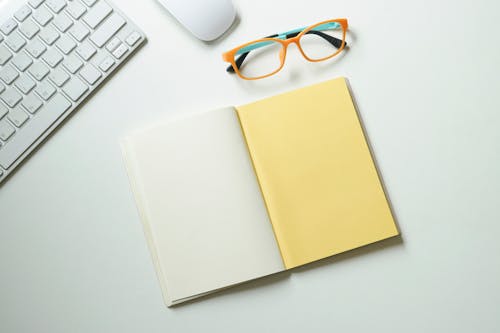 Gratis Notebook Putih Dan Kuning Ditempatkan Di Dekat Keyboard Dan Kacamata Foto Stok