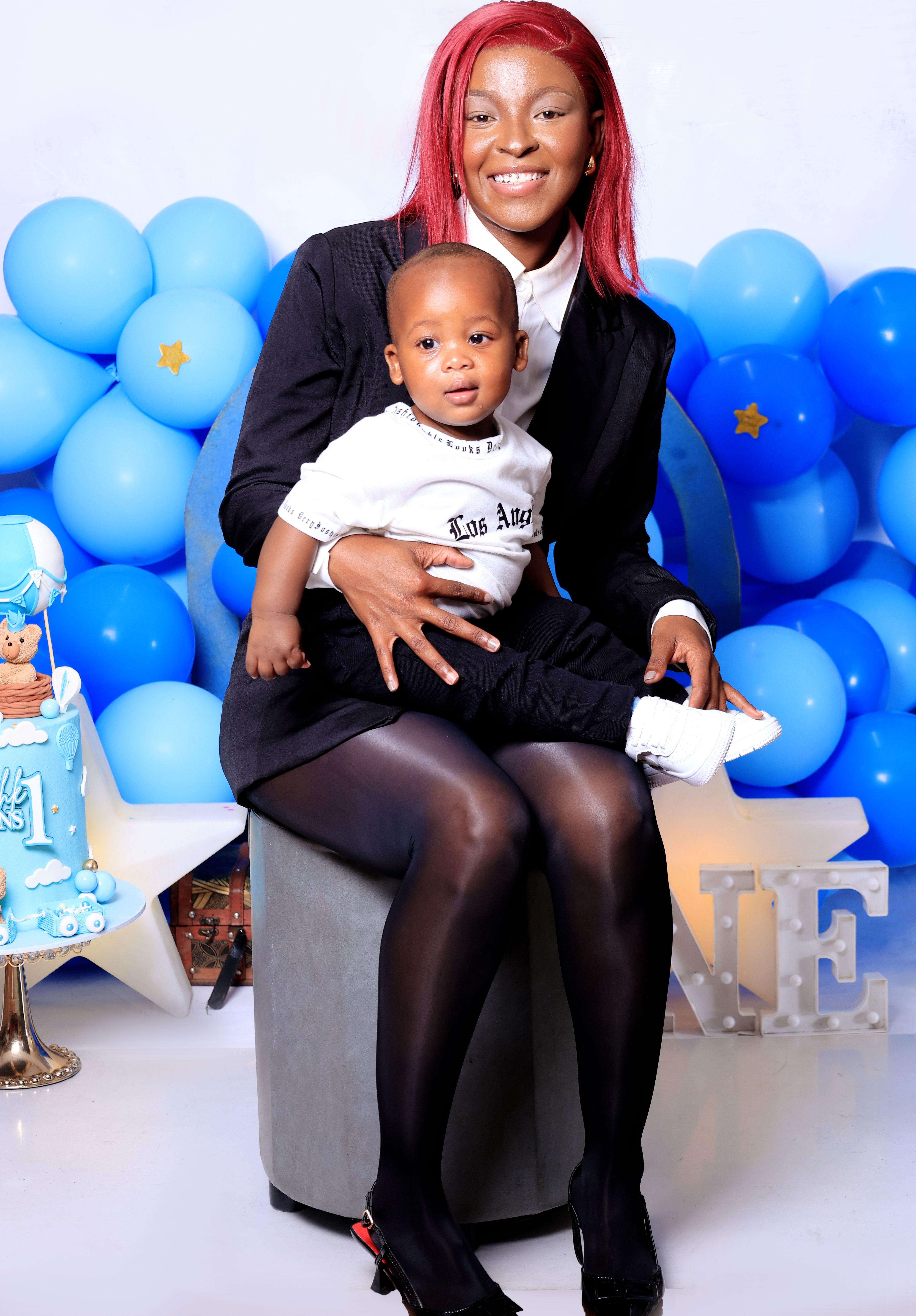 Smiling mother with red hair holds her baby during a joyful first birthday celebration with blue balloons and cake.